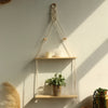 A macrame wood floating wall shelf with a hanging rope displaying candles and a potted plant on a light-colored wall.