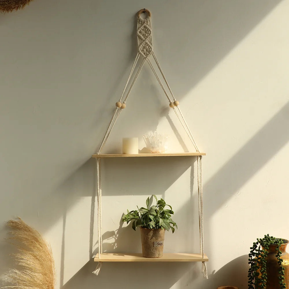 A macrame wood floating wall shelf with a hanging rope displaying candles and a potted plant on a light-colored wall.