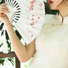 A woman holds an elegant white folding fan decorated with red plum blossoms.