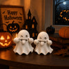 A set of two white kawaii ghost statues for Halloween decor placed on a wooden surface next to a pumpkin and a book.