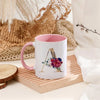 A white ceramic coffee mug with a pink interior and a gold letter initial design featuring colorful flowers.