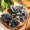 A cluster of dark gray and translucent zinc crystals resting on a wooden surface next to green leaves.