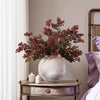 A faux branch of dusty plum nandina berries in a light gray vase on a wooden table.