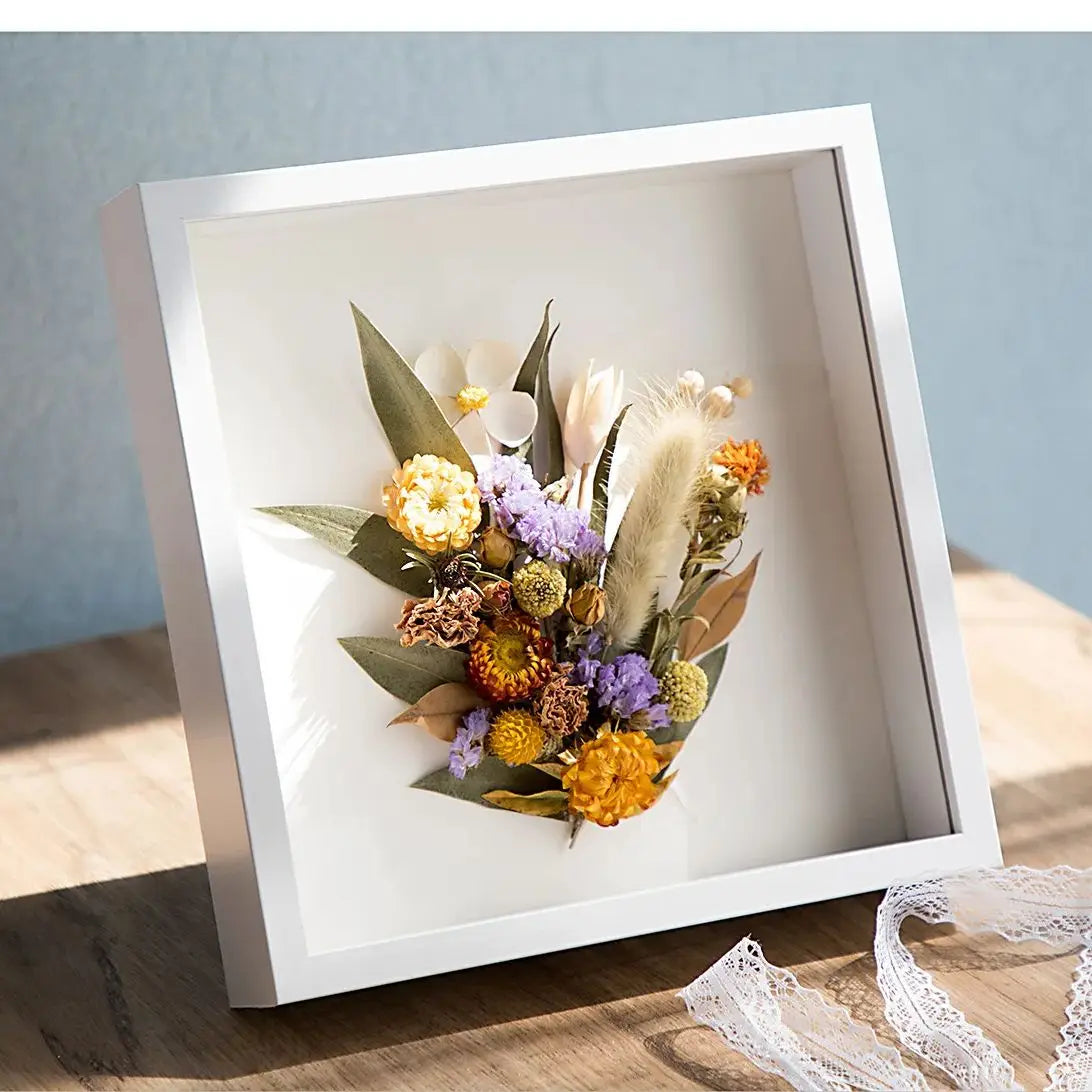 A white wooden shadow box frame containing dried floral arrangements and natural elements.