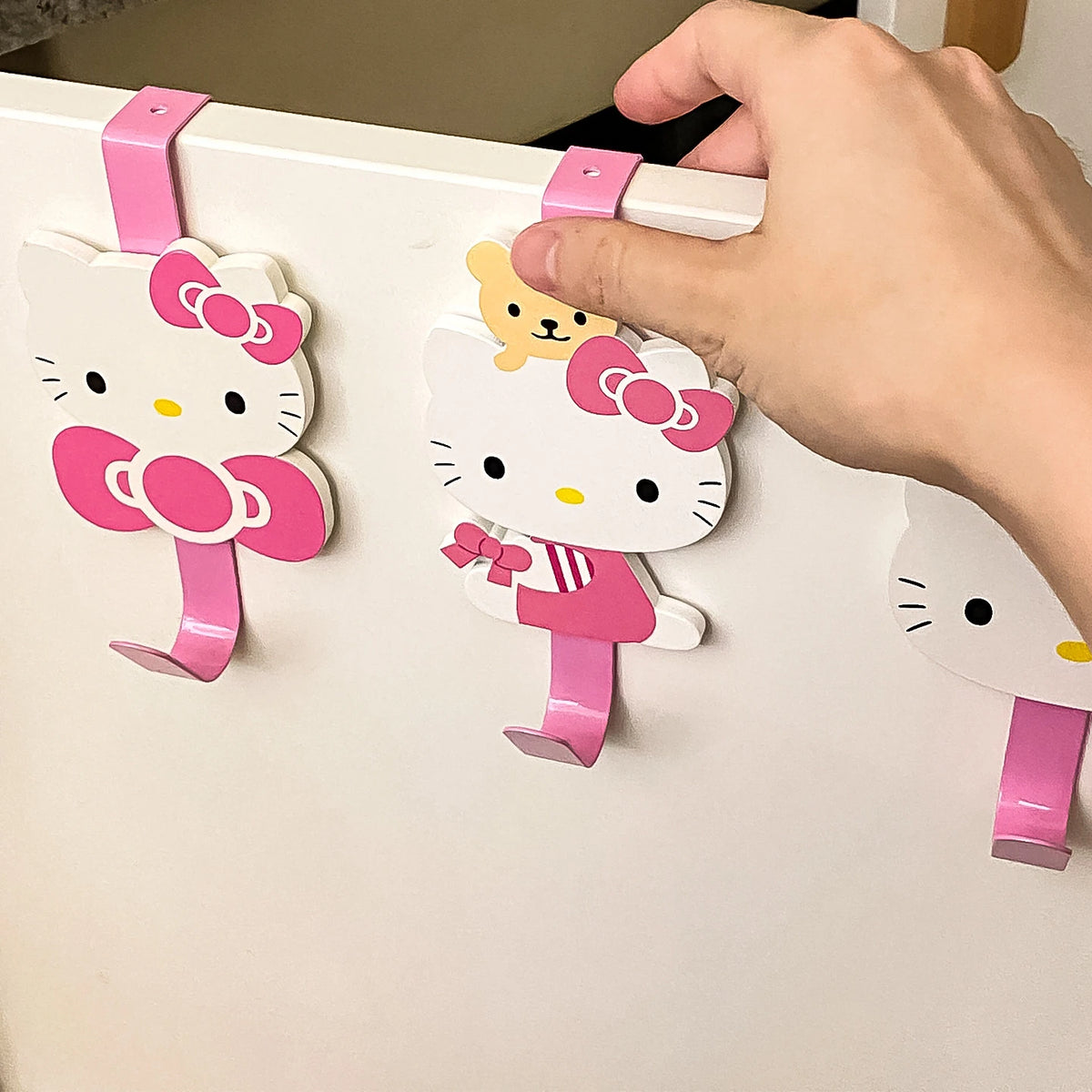 A hand is hanging a pink and white cartoon cat hook onto a white door with a pink hanger.