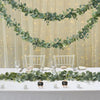 A white table decorated with a green eucalyptus vine garland and glassware, set against a sheer curtain backdrop.