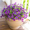 A vibrant bouquet of purple and green artificial flowers in a beige pot sitting on a wooden deck.