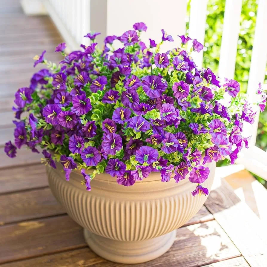 A vibrant bouquet of purple and green artificial flowers in a beige pot sitting on a wooden deck.