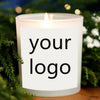 A white candle in a glass holder with a custom label reading your logo, set against green foliage.