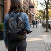 A person wearing a denim jacket and dark pants is carrying a black leather backpack with a bat charm on a city sidewalk.