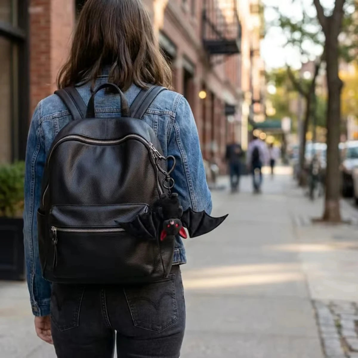 A person wearing a denim jacket and dark pants is carrying a black leather backpack with a bat charm on a city sidewalk.