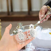 A hand holds a decorative glass bottle with a silver figurine and twist cap on a table.