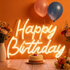 An illuminated neon wall sign spelling out Happy Birthday with a cake in front of it.