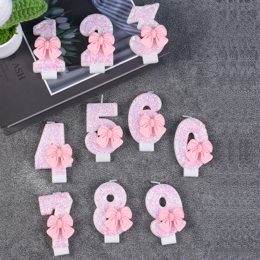 A collection of glitter number candles from 1 to 9, featuring pink bows, displayed on a dark gray surface.