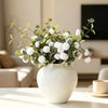 A rustic textured round white vase filled with green and white foliage sits on a light-colored table.