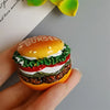 A hand holds a burger shaped food magnet with lettuce, tomato, and cheese layers.