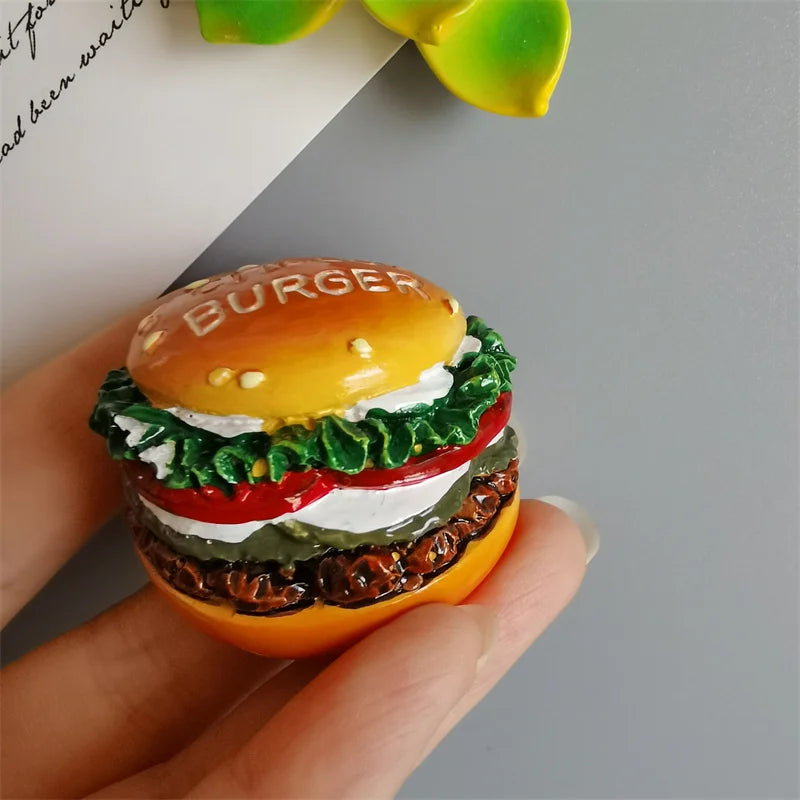 A hand holds a burger shaped food magnet with lettuce, tomato, and cheese layers.