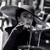 A woman wearing a wide-brimmed hat and black clothing is dipping a finger into a bowl of food.