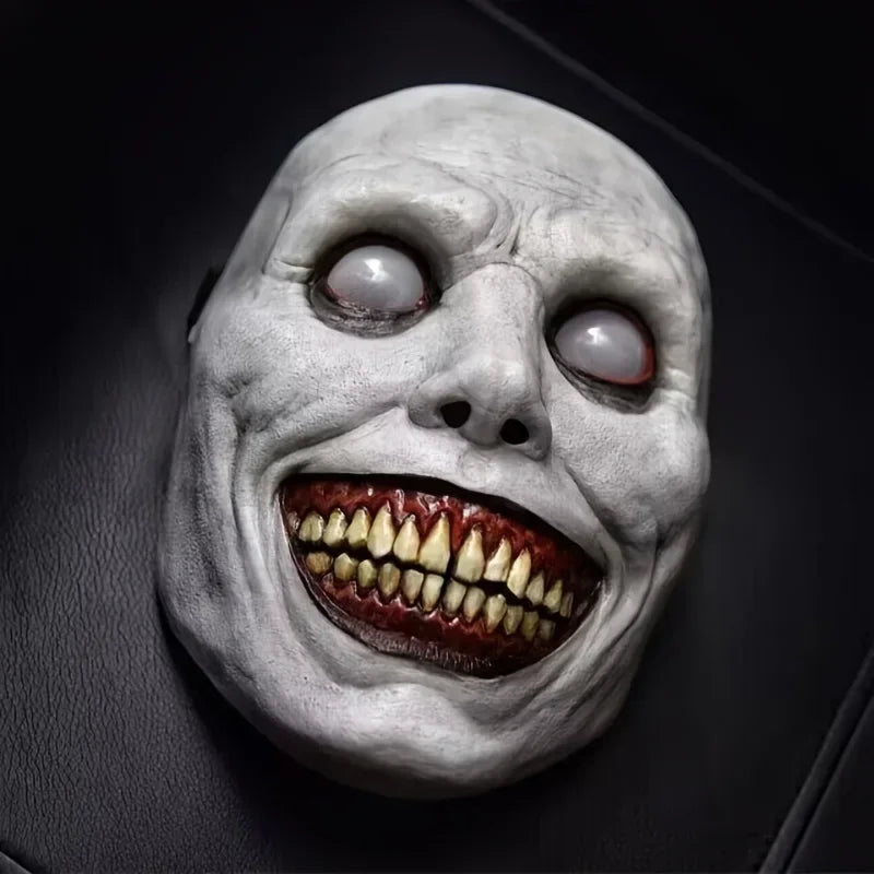 A realistic smiling demon latex mask with pale skin, glowing eyes, and a wide, toothy grin.