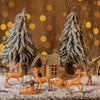 A set of mini deer figurines in various colors standing in a snowy outdoor scene with evergreen trees and a small wooden hous