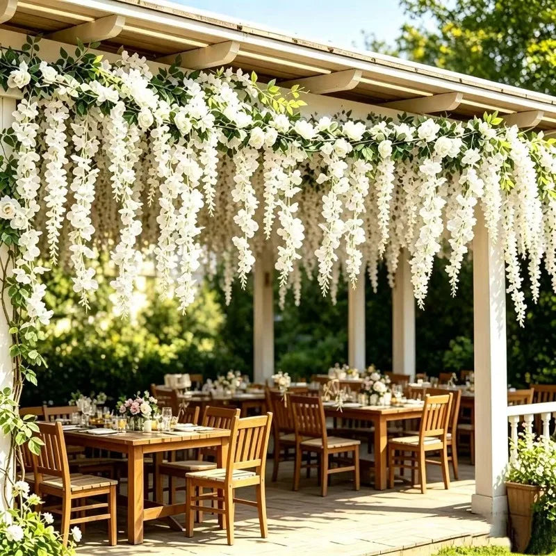 A wooden outdoor dining area decorated with white silk wisteria hanging vine garlands and greenery.