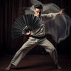 A male model in a white gi performs a martial arts pose while holding a black folding hand fan.