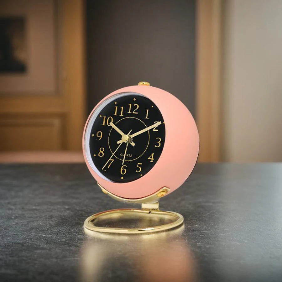 A pink minimalist Nordic alarm clock with a black face and gold accents sitting on a dark surface.