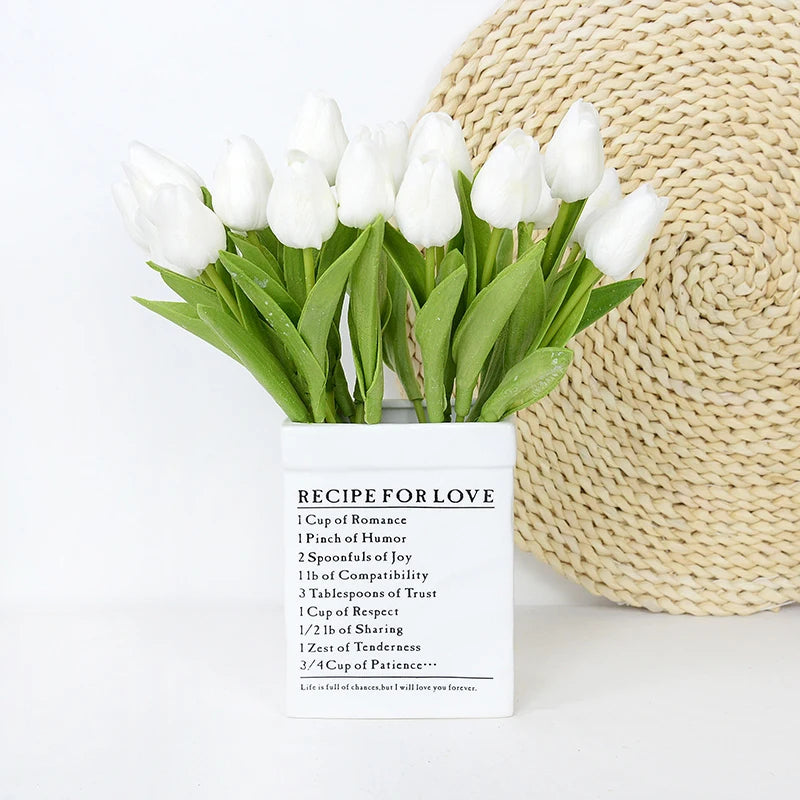 A bouquet of white artificial tulips in a square white container next to a woven beige sphere.