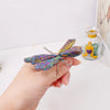 A hand holds a natural crystal tourmaline dragonfly figurine with iridescent blue and purple wings.