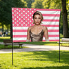A woman wearing a sheer top stands in front of a pink and white American flag banner in a park setting.
