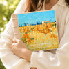 A hand holds a laminated photo album featuring an oil painting of a golden field under a blue sky.