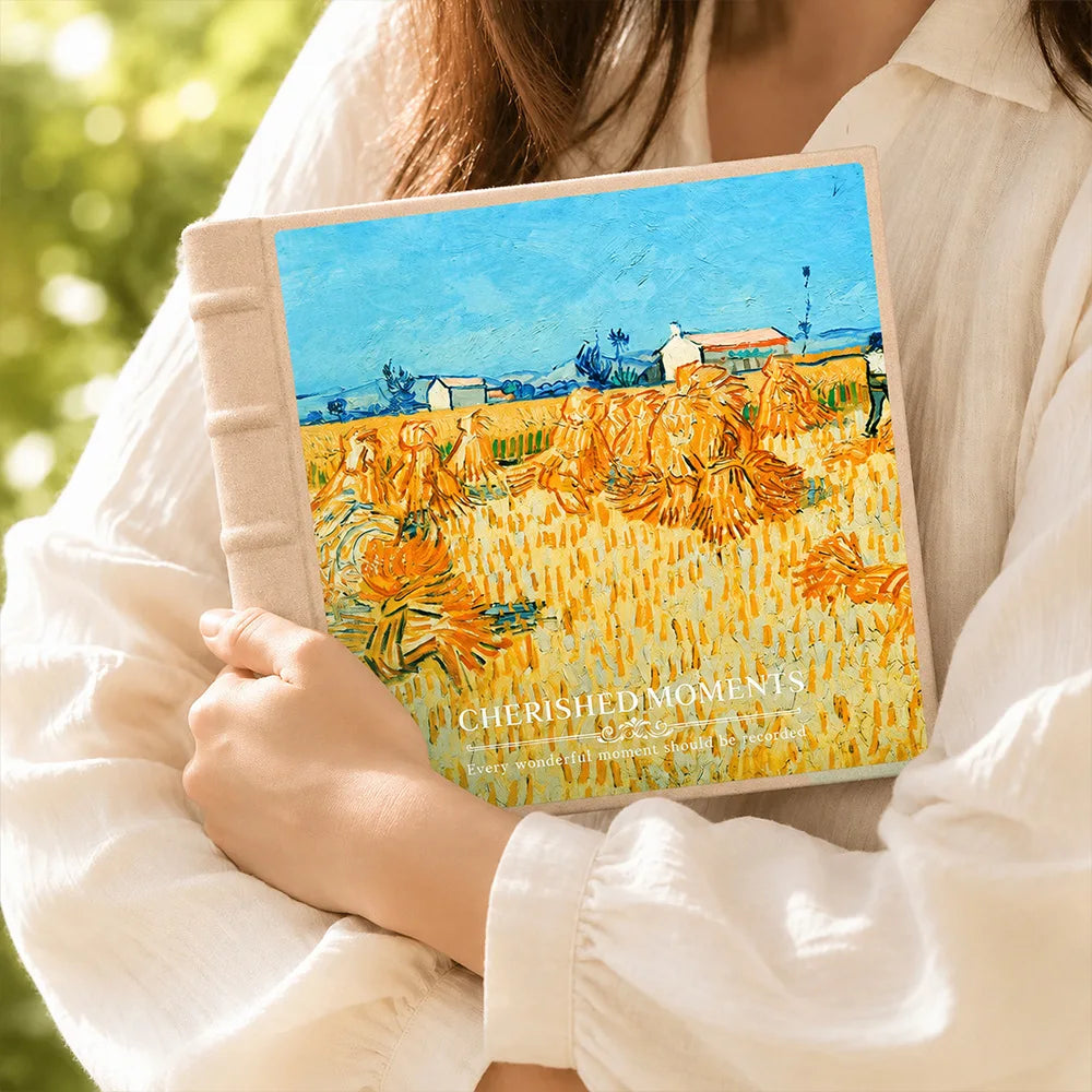 A hand holds a laminated photo album featuring an oil painting of a golden field under a blue sky.