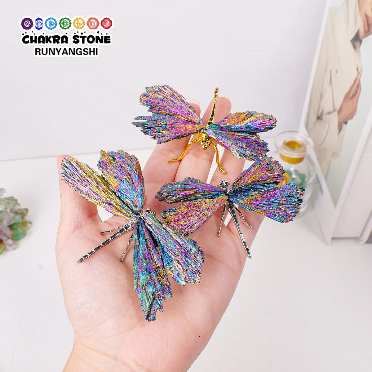 A hand holds a sparkling crystal tourmaline dragonfly figurine with iridescent purple and blue wings.