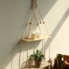 A macrame wood floating wall shelf with hanging rope displaying a candle and a potted plant on a light-colored wall.