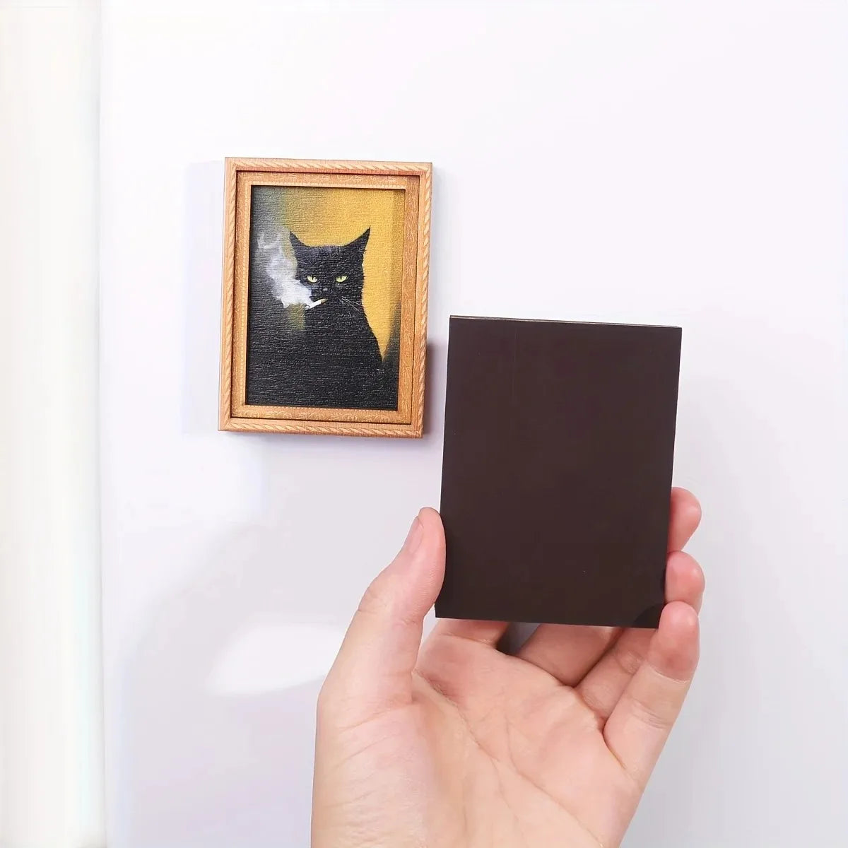 A hand holds a dark brown refrigerator magnet framed in wood, featuring a silhouette of a black cat against a yellow backgrou