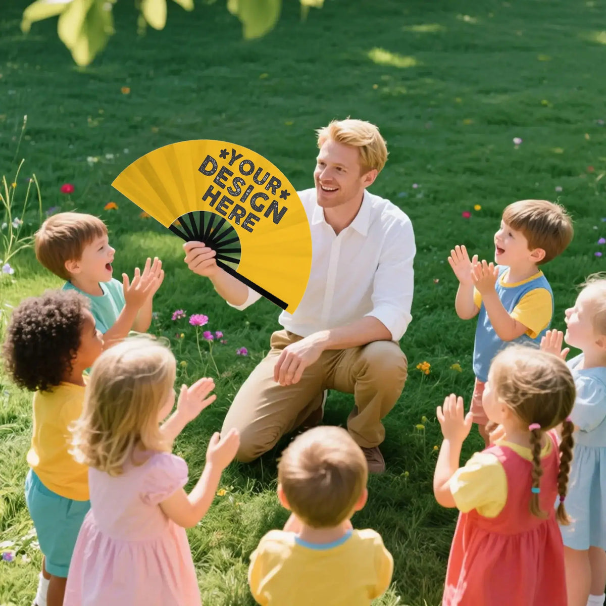 A young man holds a yellow folding hand fan while several children look on in a grassy outdoor setting.