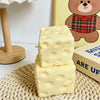 A stack of yellow cheese squishy stress relief toys resting on a woven mat next to a teddy bear illustration.