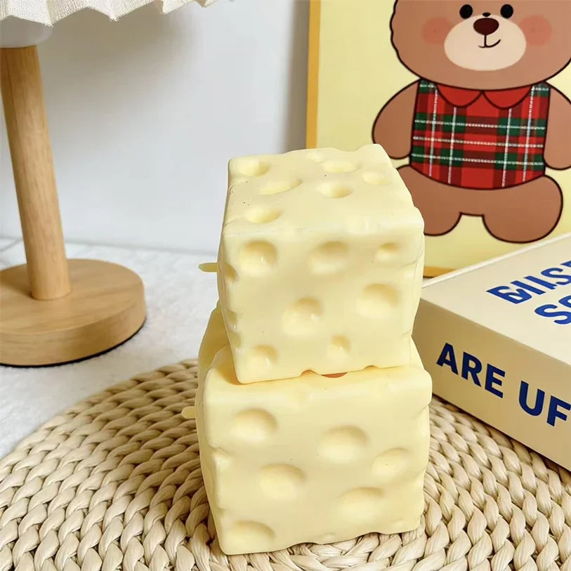 A stack of yellow cheese squishy stress relief toys resting on a woven mat next to a teddy bear illustration.