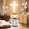 A crochet doll with a smiling face and an encouragement card sits in front of a glowing cross and a dove, set on a wooden sur