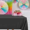 A pink and blue birthday cake with candles sits on a black tablecloth in front of a white brick wall.