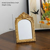 A gold-framed arched carved mirror with a rectangular reflective surface displayed on a wooden surface.