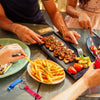 Hands are preparing food on a wooden table, featuring french fries and meat skewers, with an aluminum bottle opener keychain 