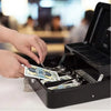 A hand is placing US dollar bills into a black lockable metal cash box with an organizer tray and two keys.