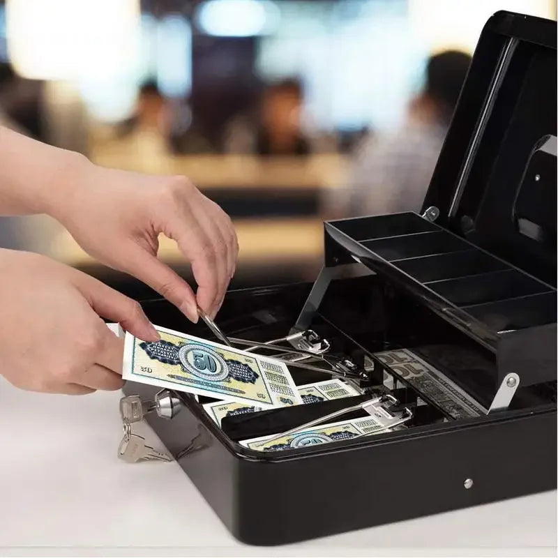 A hand is placing US dollar bills into a black lockable metal cash box with an organizer tray and two keys.