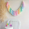 Handmade boho rainbow tassel garland with wooden beads hanging on a white wall above a small cake.