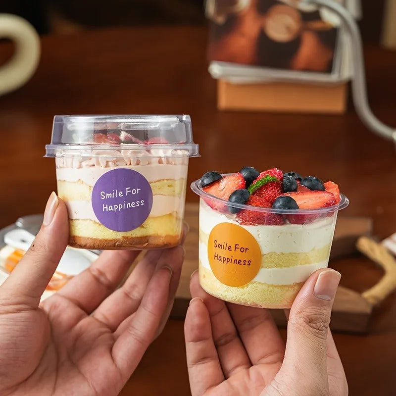Two clear plastic pudding cups filled with layered dessert are being held by a hand on a wooden table.