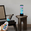 A hand holds a remote control next to a tall, cylindrical LED fish lamp sitting on a wooden table.