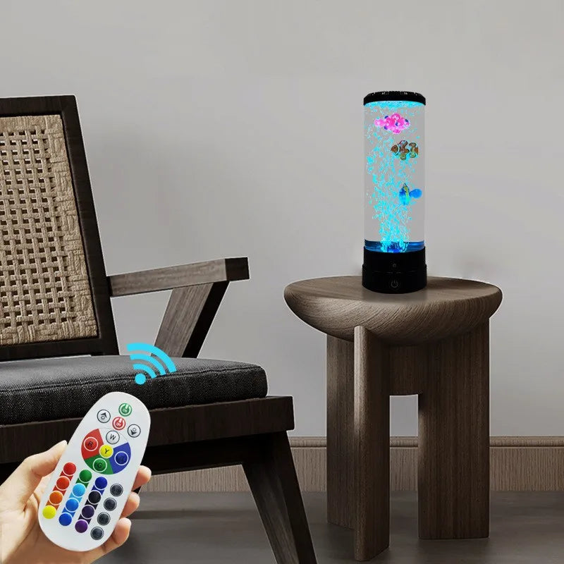 A hand holds a remote control next to a tall, cylindrical LED fish lamp sitting on a wooden table.