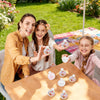 Three children are shown making DIY stress balls with filling beads on a wooden table outdoors.