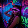A woman wearing neon purple light up wire party glasses posing against a colorful graffiti wall.